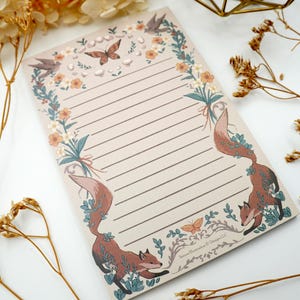 May include: A rectangular notepad with horizontal lines, featuring a decorative border of orange flowers, green foliage, two brown foxes, a butterfly, and birds. The bottom right corner displays the text Satyro Illustration & Design LLC.