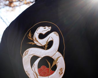 May include: Black hoodie with a white embroidered snake design on the back. The snake is coiled around a red and gold floral pattern.