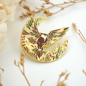 May include: A gold-colored pin featuring an eagle with outstretched wings, set against a crescent moon shape. The eagle's feathers are detailed with shades of brown, green, and tan. The pin is surrounded by leaf-like details.