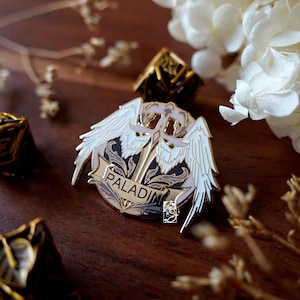 May include: A detailed Paladin enamel pin with white wings, a sword, and the word "PALADIN" on a banner. The pin is set against a dark wooden surface with decorative elements and a gold-colored die.