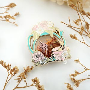 May include: A decorative enamel pin featuring a teapot design with a light blue outline, pink roses, and a teacup. The pin has a clear center with a glittery, brown background and a pink cat illustration.