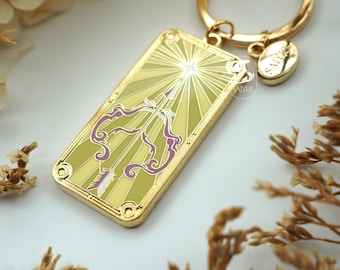 Könnte beinhalten: Ein rechteckiger Schlüsselanhänger aus goldfarbenem Metall mit einer stilisierten violetten Pfeil-und-Bogen-Grafik vor einem strahlenden hellgelben Hintergrund. Das Design enthält einen kleinen runden Anhänger mit dem Text SAIYRI am goldenen Ring.