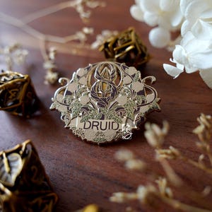 May include: A gold-colored Druid enamel pin with floral and leaf details. The pin features the word "DRUID" in a banner across the bottom. The pin is surrounded by other decorative items, including dice and dried flowers, on a wooden surface.