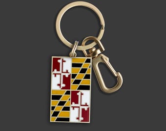 May include: Gold-coloured keychain with a rectangular charm displaying the Maryland state flag. The flag design features red, white, black, and yellow geometric shapes and crosses. The keychain has a ring and a clasp.