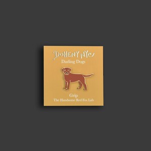 May include: A gold pin with a brown Labrador Retriever silhouette. The pin says "Doheny NYC" and "Darling Dogs" above the dog and "Grip" and "The Handsome Red Fox Lab" below the dog.