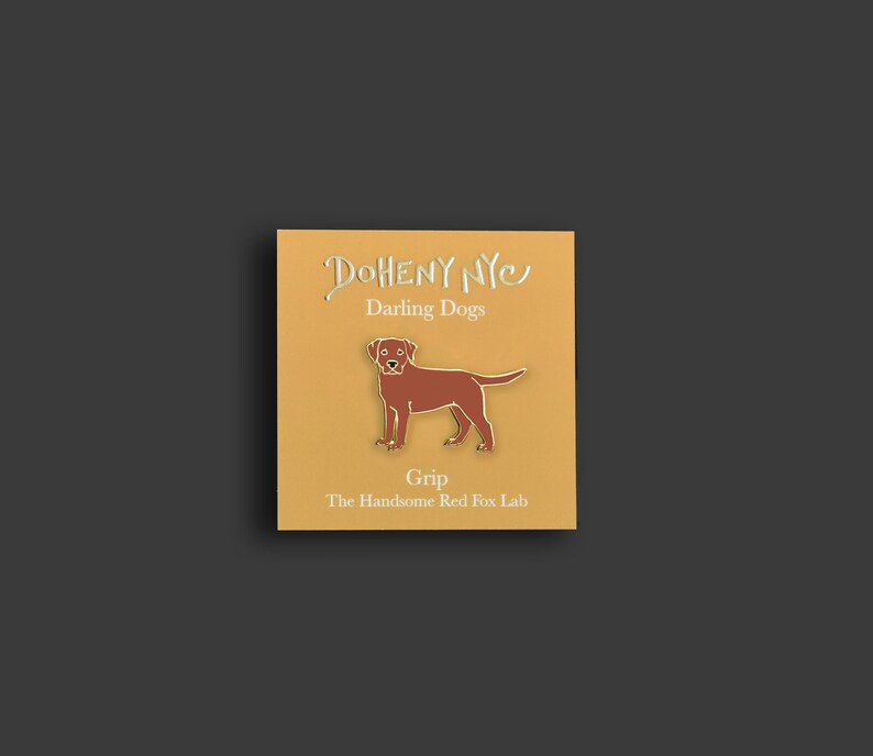 May include: A gold pin with a brown Labrador Retriever silhouette. The pin says "Doheny NYC" and "Darling Dogs" above the dog and "Grip" and "The Handsome Red Fox Lab" below the dog.