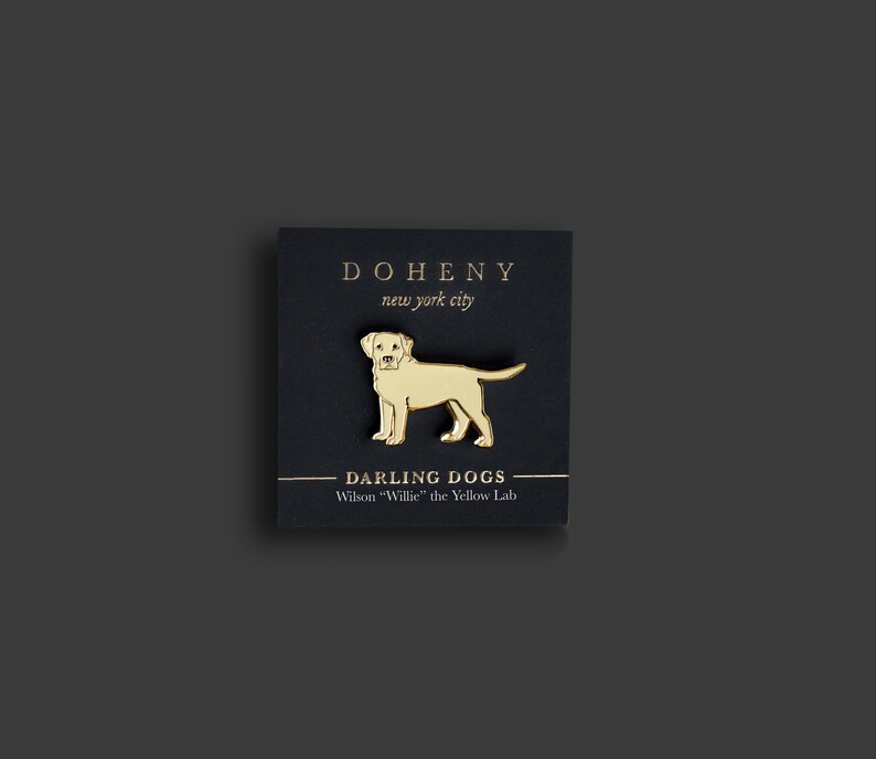 Op de afbeelding: Een goudkleurige pin van een Labrador Retriever-hond, met het label "Wilson 'Willie' the Yellow Lab", wordt getoond op een zwarte kaart. De kaart bevat ook de tekst "DOHENY new york city" in gouden letters. De pin is een gedetailleerde illustratie van de hond.