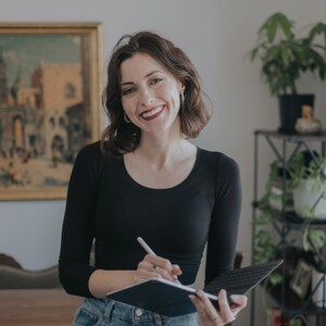 Op de afbeelding: Een vrouw in een zwart shirt en een blauwe jeans lacht terwijl ze een tablet en stylus vasthoudt. Een schilderij hangt aan de muur achter haar. Potplanten zijn zichtbaar op de achtergrond.