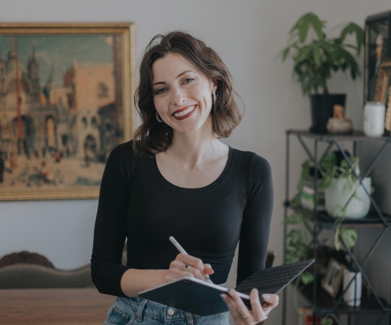 Op de afbeelding: Een vrouw in een zwart shirt en een blauwe jeans lacht terwijl ze een tablet en stylus vasthoudt. Een schilderij hangt aan de muur achter haar. Potplanten zijn zichtbaar op de achtergrond.