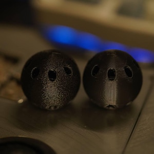 Puede incluir: Dos objetos esféricos negros con tres agujeros ovalados. Los objetos tienen una superficie texturizada y parecen impresos en 3D. El fondo está borroso, con un toque de luz azul. Medidas en centímetros.
