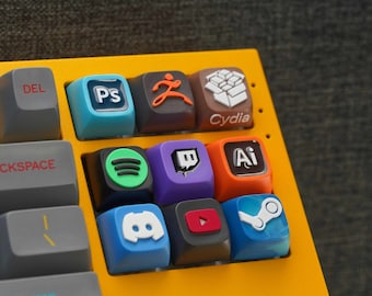 May include: A yellow keyboard with custom keycaps featuring various logos, including Spotify, Twitch, Discord, and Adobe Illustrator. The keycaps are colorful and have a glossy finish.