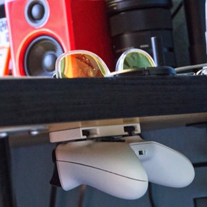 Op de afbeelding: Twee witte videogame controllers gemonteerd onder een zwart rek met een zonnebril op het rek.