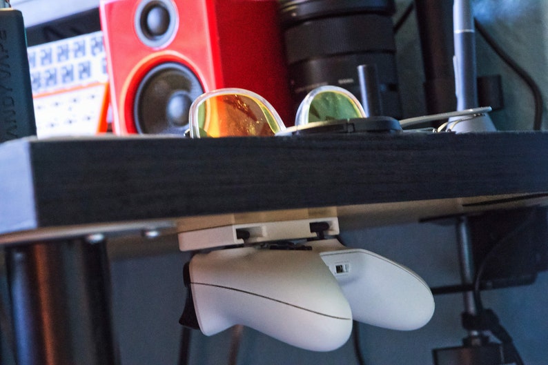 Op de afbeelding: Twee witte videogame controllers gemonteerd onder een zwart rek met een zonnebril op het rek.