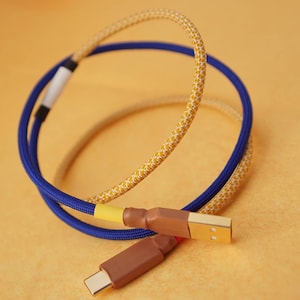 May include: A close-up of a blue and yellow braided cable with a gold-colored USB-C connector. The cable is wrapped in a braided fabric with a yellow and white pattern.