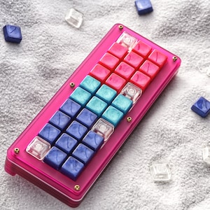 May include: A pink, blue, and red mechanical keyboard with a clear plastic case. The keyboard has a unique layout with a variety of keycaps.