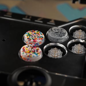 Puede incluir: Primer plano de un mando de arcade negro con botones coloridos. Dos botones están cubiertos de chispas de arcoíris, uno es gris con un diseño de mármol. El mando tiene interruptores mecánicos transparentes.