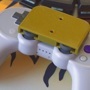 Op de afbeelding: Een witte videogamecontroller met een gele 3D-geprinte bevestiging erop. De controller heeft vier paarse knoppen en &eacute;&eacute;n grijze knop.