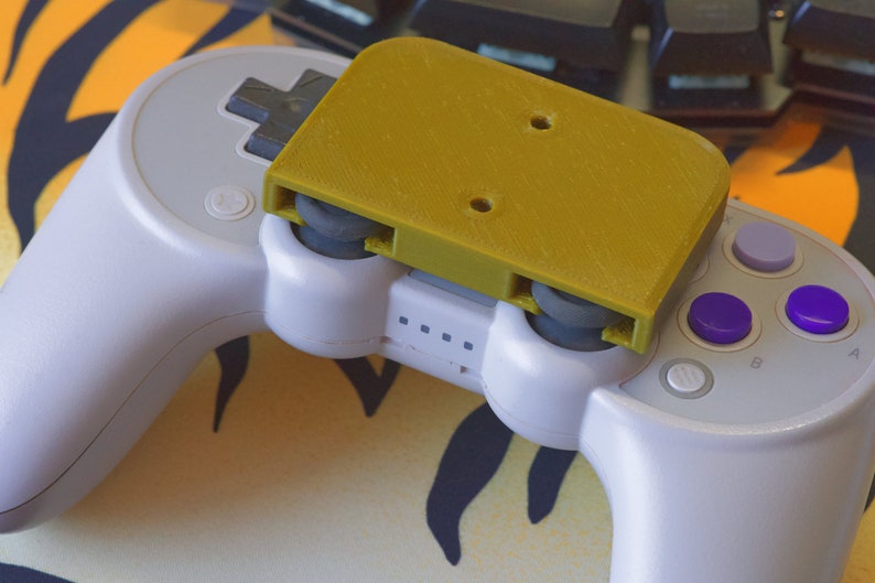 Op de afbeelding: Een witte videogamecontroller met een gele 3D-geprinte bevestiging erop. De controller heeft vier paarse knoppen en &eacute;&eacute;n grijze knop.