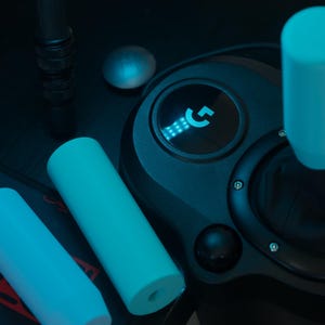 Puede incluir: Un volante de juego negro con un logotipo G azul iluminado y una perilla de cambio de marchas azul. El volante tiene una superficie texturizada y un diseño circular.