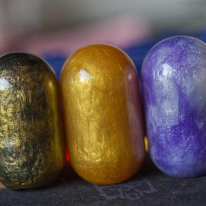 Puede incluir: Tres objetos redondos, lisos, parecidos a la resina con un acabado brillante. Los objetos están en fila de izquierda a derecha: negro con motas doradas, dorado con un ligero brillo y morado con motas blancas.