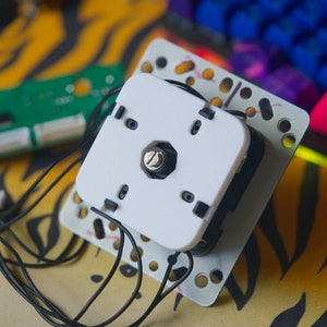 Może przedstawiać: Biały joystick z czarnym pokrętłem i srebrną śrubą. Joystick jest podłączony do płytki drukowanej czarnymi przewodami.