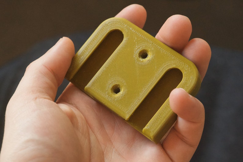 Op de afbeelding: Een 3D-geprint clip van olijfgroen plastic met twee gleuven en twee gaten.