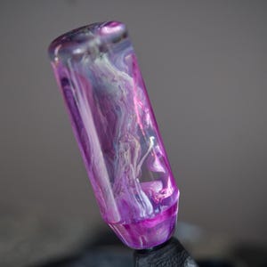 Peut inclure: Un pommeau de levier de vitesse en résine transparente, de couleur violette et blanche, avec des motifs tourbillonnants.