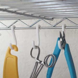 Op de afbeelding: Een metalen draadplank met witte plastic haken die diverse gereedschappen vasthouden. Een gele snijplank, zilveren schaar en blauwe tang hangen aan de haken, wat een opbergsysteem laat zien.