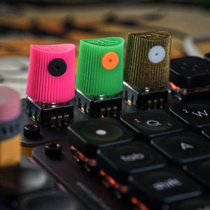 Puede incluir: Tres botones coloridos con tapas de diferentes colores, rosa, verde y marrón, están montados en una superficie negra. Los botones están conectados a un teclado negro con teclas blancas.