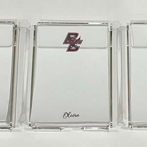 May include: Three clear acrylic notepads with personalised names and college logos. The first notepad has a green "O" for Oregon and the name "Maya". The second notepad has a maroon "BC" for Boston College and the name "Olivia". The third notepad has a green "V" for Villanova and the name "Sadie".