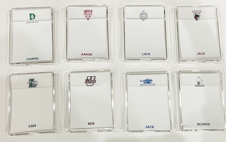 May include: Eight clear acrylic notepads with college logos and names. The logos include Dartmouth, WPI, a crest, Tartans, UMass, Middlebury, a crest, and Bowdoin. The names are Cooper, Aaron, Luca, Jack, Sam, Ben, Jack, and Reuben.