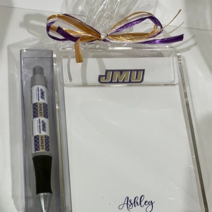 Papel de notas con temática universitaria en un soporte de metacrilato y un bolígrafo a juego.