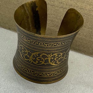 May include: A wide, open cuff bracelet in a brassy gold color. The cuff features an intricate, repeating pattern of swirling designs and lines. The bracelet has a slightly curved shape and is open on the top and bottom.