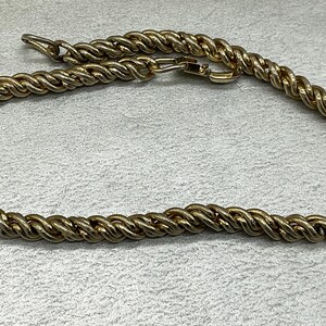 May include: A gold-colored, twisted rope chain necklace. The necklace is made of a series of intertwined strands, creating a textured, rope-like appearance. The clasp is visible, and the necklace is lying on a light gray surface.