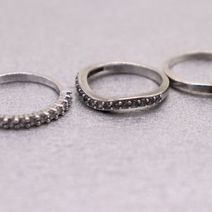 May include: Three silver rings with small clear stones. The ring on the left has a row of stones around the band. The middle ring has a curved band with stones around the edge. The ring on the right has a single stone on the top.