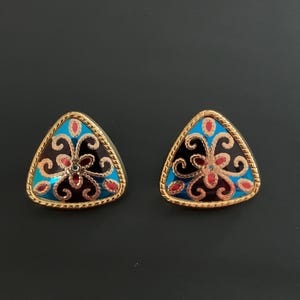 May include: A pair of triangular earrings featuring a gold-toned rope-textured border. The center displays a vibrant enamel design with a black background, bright blue accents, and small red teardrop-shaped details arranged in a symmetrical pattern.