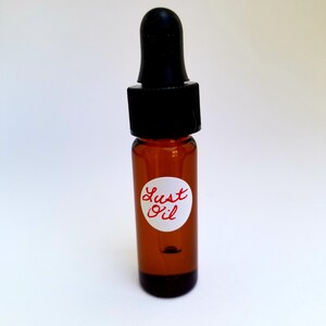 May include: A small brown glass bottle with a black dropper top. The bottle has a white label with the words "Lust Oil" written in red.