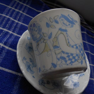 Może przedstawiać: Biały kubek do herbaty i spodek z niebieskim i białym wzorem kwiatowym. Kubek do herbaty przedstawia fantazyjną ilustrację dwóch postaci w stylizowanym wzorze.