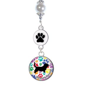 May include: A black braided leather bracelet with a silver clasp and a charm featuring a black paw print and a colourful Basset Hound design. The charm has a white pearl and silver accents.