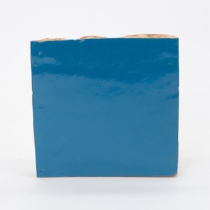 Puede incluir: Un azulejo cuadrado con un acabado brillante de color azul intenso. El azulejo tiene un borde áspero y desigual.
