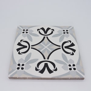 May include: A white ceramic tile with a black and gray geometric pattern. The pattern features stylized floral motifs and a central diamond shape.