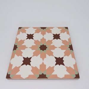 May include: A square ceramic tile with a geometric pattern in shades of peach, white, brown, and green. The pattern features a repeating design of stars and interlocking shapes.