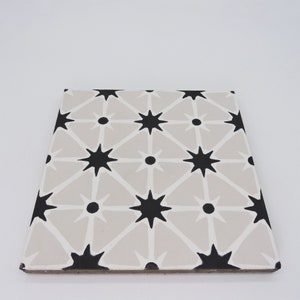 May include: A square ceramic tile with a geometric pattern of black stars and white lines on a light gray background.