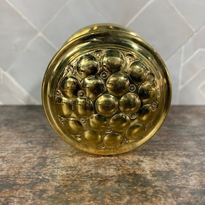 May include: A gold-toned metal container with a round, embossed lid. The lid features a repeating pattern of raised circles.