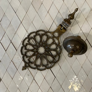 May include: A brass decorative door knocker with a circular, intricate design and a domed top. The knocker is attached to a white tiled wall.
