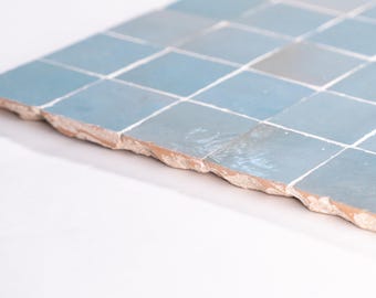 May include: A section of square, light blue tiles with white grout lines. The tiles have a slightly uneven, textured surface and a rough, exposed edge. The color is a muted, cool tone, suitable for home decor.