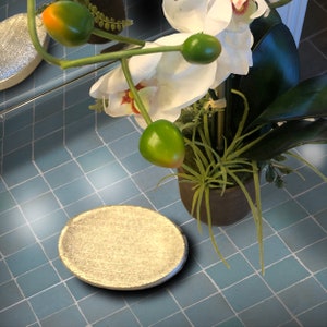 Könnte beinhalten: Eine runde, metallisch goldfarbene Seifenschale mit strukturierter Oberfläche. Die Schale steht auf einer blau gefliesten Oberfläche, mit einer weißen Orchidee und grünen Akzenten im Hintergrund.