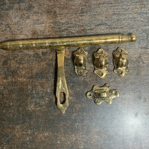 May include: A gold-toned metal door latch with a decorative design. The latch has a long, straight bar with a handle on one end and a hook on the other. There are four smaller decorative pieces with intricate designs.