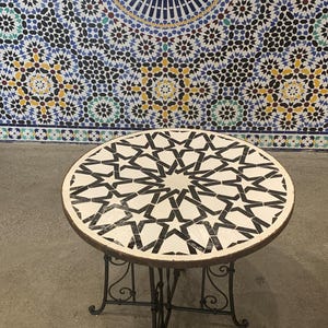 Puede incluir: Mesa redonda de mosaico con un diseño de estrella en blanco y negro. La mesa tiene una base de metal oscuro con adornos decorativos. El fondo presenta una colorida pared de mosaicos con diseños geométricos.