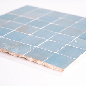 Könnte beinhalten: Ein Abschnitt quadratischer, hellblauer Fliesen mit weißen Fugen. Die Fliesen haben eine leicht unebene Oberfläche und ein rustikales, handgefertigtes Aussehen. Die Kanten des Fliesenabschnitts sind rau und unfertig und zeigen eine terrakottafarbene Basis.